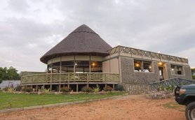 Ein modernes Safari-Lodge-Gebäude mit einem markanten, strohgedeckten Dach und einer einladenden Veranda umgeben von Pflanzen.