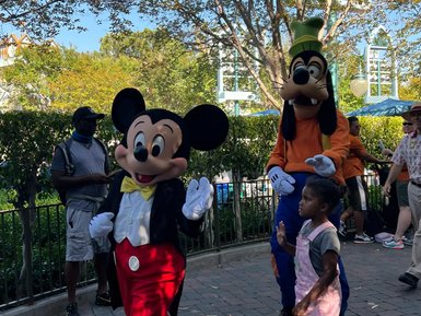 Mickey Mouse und Goofy gehen fröhlich durch einen Park, während ein kleines Mädchen ihnen neugierig folgt.