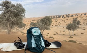 Ein Rucksack und ein Snowboard liegen auf einer Sanddüne, umgeben von vereinzelten Bäumen in einer Wüstenlandschaft.