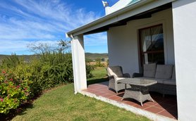 Eine gemütliche Terrasse mit einem Rattansofa und einem Tisch, umgeben von üppigem Grün und blühenden Pflanzen.
