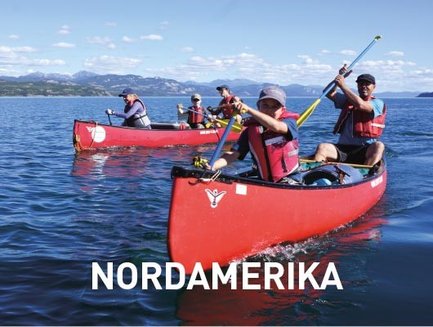 Vier Kanuten in roten Booten paddeln auf einem ruhigen Gewässer, umgeben von Bergen und einem klaren blauen Himmel.