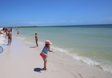 Ein sonniger Strand mit Familien, die im Wasser spielen und am Ufer entspannen, während die Wellen sanft an den Sand schlagen.