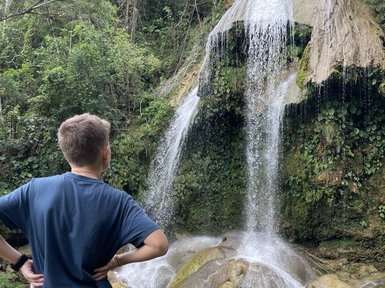 Ein Junge steht am Ufer eines klaren Wassers, während ein beeindruckender Wasserfall hinter ihm herabstürzt.