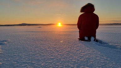 Eine Person in einem roten Mantel kniet im Schnee und beobachtet den Sonnenuntergang über einer verschneiten Landschaft.