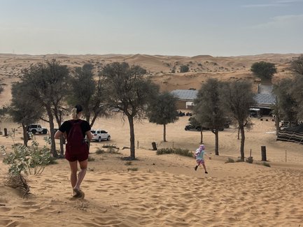 Eine Person mit Rucksack wandert eine sandige Düne hinunter, während ein Kind fröhlich im Hintergrund spielt.