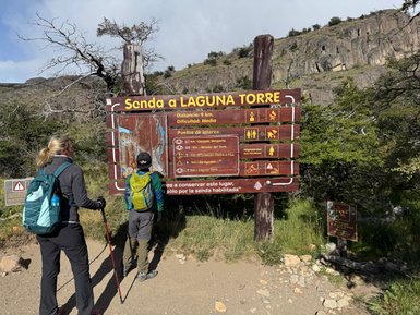 Zwei Wanderer betrachten ein Informationsschild, das den Weg zur Laguna Torre in Patagonien beschreibt, umgeben von Natur.