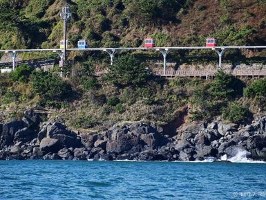 Eine farbenfrohe Seilbahn fährt über eine grüne Küstenlandschaft, während Wellen sanft gegen die Felsen schlagen.