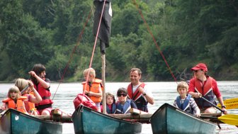 Eine Gruppe von Kindern und Erwachsenen paddelt in Kanus auf einem ruhigen Gewässer, umgeben von üppigem Grün.