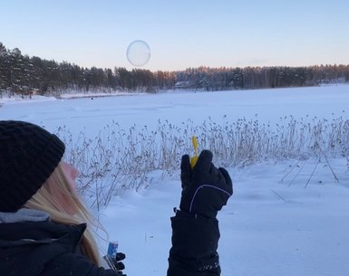 Eine Person in warmer Kleidung bläst Seifenblasen in einer verschneiten Landschaft, während die Sonne am Horizont aufgeht.