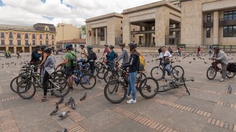 Eine Gruppe von Radfahrern steht auf einem Platz in Bogotá, umgeben von Tauben und historischen Gebäuden.