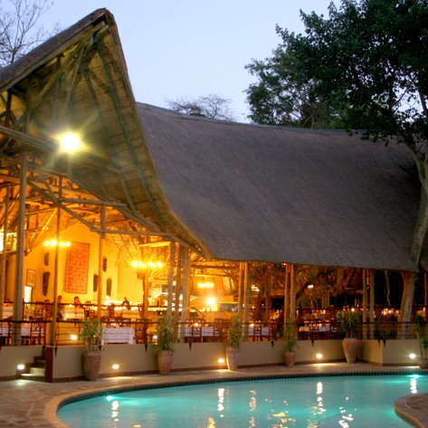 Ein stilvolles Safari-Lodge mit einem großen Strohdach, beleuchtet von warmem Licht, umgeben von Bäumen und einem Pool.