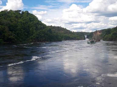 Ein Boot fährt auf einem ruhigen Fluss, umgeben von üppigem Grün und sanften Hügeln, mit einem Wasserfall im Hintergrund.