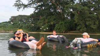 Fünf Personen entspannen auf Reifen im Wasser, umgeben von üppigem Grün und fliegenden Vögeln im Hintergrund.