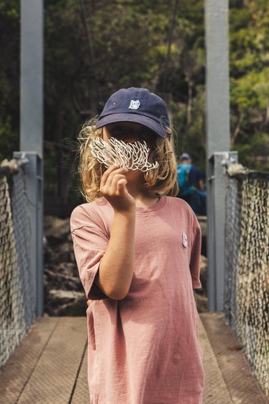 Ein Kind mit blonden Locken hält ein Stück Koralle vor dem Gesicht, während es auf einer Hängebrücke steht.