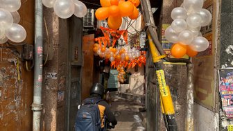 Ein Motorradfahrer fährt durch eine enge Gasse, geschmückt mit orangefarbenen und transparenten Luftballons.