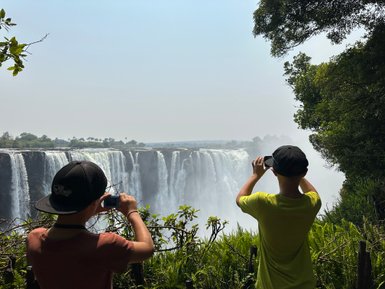Zwei Kinder stehen am Rand eines Wasserfalls und fotografieren die beeindruckende Aussicht auf die Victoriafälle.