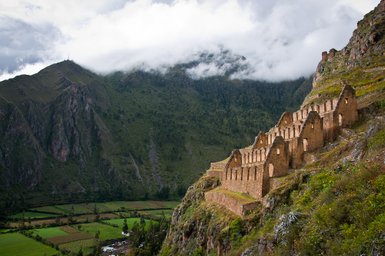 Die Ruinen von Ollantaytambo thronen majestätisch auf einem Berghang, umgeben von üppigen grünen Feldern und dramatischen Bergen.