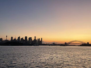 Der lebhafte Sonnenuntergang über Sydney malt die Skyline in warme Gold- und Orangetöne, während das Wasser glitzert.