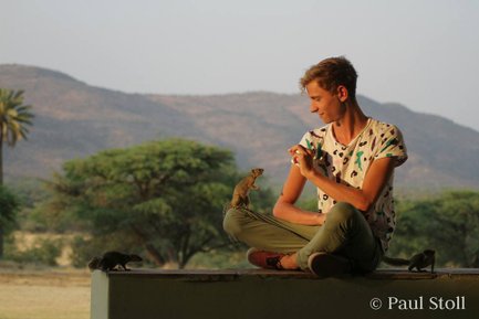 Ein junger Mann sitzt entspannt auf einer Mauer und interagiert freundlich mit einem kleinen Tier in der Natur.