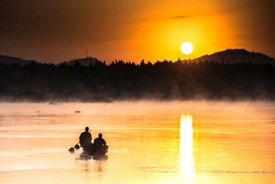 Zwei Personen paddeln in einem Boot über einen ruhigen See, während die Sonne hinter den Bergen untergeht.