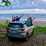 Eine Gruppe von vier Personen steht fröhlich neben einem silbernen Auto am Strand von Costa Rica, umgeben von üppigem Grün.
