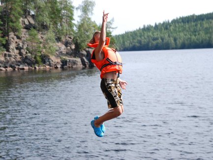 Ein Junge springt fröhlich ins Wasser, trägt eine orangefarbene Schwimmweste und blaue Crocs, umgeben von Natur.