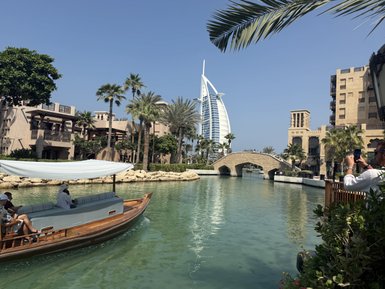 Ein traditionelles Boot fährt durch eine grüne Wasserstraße, umgeben von modernen und historischen Gebäuden in Dubai.