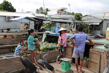 Auf einem Markt im Mekong-Delta stehen Menschen umgeben von Wassermelonen, während ein Hund im Hintergrund sitzt.