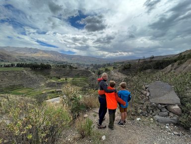 Eine Frau mit zwei Kindern steht an einem Hang und bewundert die terrassierten Felder im Colca-Canyon unter einem bewölkten Himmel.