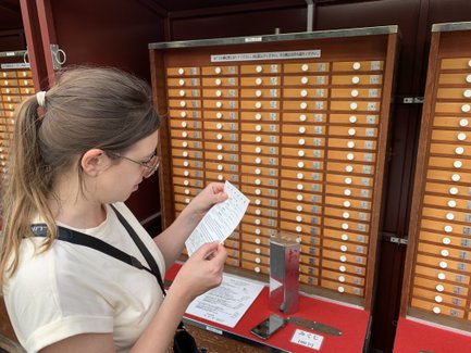 Eine Person mit Brille liest einen Zettel vor einer Wand mit vielen kleinen Schubladen in einem Tempel.