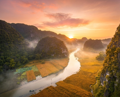 Ein atemberaubender Sonnenaufgang über einer ruhigen Flusslandschaft, umgeben von grünen Bergen und goldenen Reisfeldern.