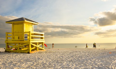 Ein leuchtend gelbes Rettungshaus steht am Strand, während Menschen im warmen Sonnenlicht spazieren gehen.