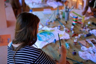 Eine Person mit langen Haaren malt konzentriert auf ein T-Shirt, umgeben von Farben und Malutensilien auf einem Holztisch.