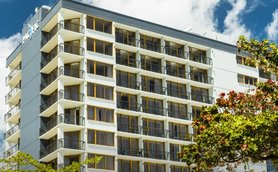 Modernes Hotelgebäude mit vielen Balkonen, umgeben von Bäumen und einem strahlend blauen Himmel.