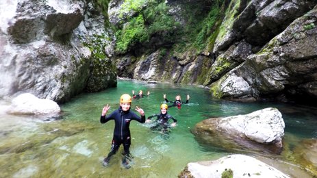 Eine Gruppe von Menschen in Neoprenanzügen genießt das Wasser eines klaren Flusses, umgeben von hohen Felsen und Bäumen.