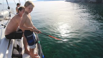 Ein Junge steht auf einem Boot und angelt, während ein Mann hinter ihm sitzt und die ruhige Wasseroberfläche betrachtet.