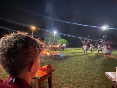 Ein Junge beobachtet eine traditionelle Tanzaufführung am Lagerfeuer, während Zuschauer im Hintergrund sitzen.