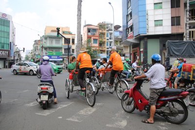 Zwei Radfahrer in orangefarbenen T-Shirts transportieren Passagiere in einem belebten Straßenverkehr in Saigon.
