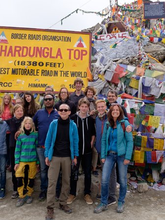 Eine Gruppe von Reisenden posiert fröhlich vor dem Khardungla-Pass-Schild, umgeben von bunten Gebetsfahnen.