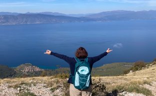 Eine Person mit einem Rucksack steht auf einem Hügel und breitet die Arme weit aus, während sie den Blick auf den Ohridsee genießt.