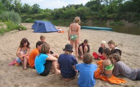 Eine Gruppe von Kindern sitzt im Sand am Flussufer, während sie an einer Campbesprechung teilnehmen und sich unterhalten.