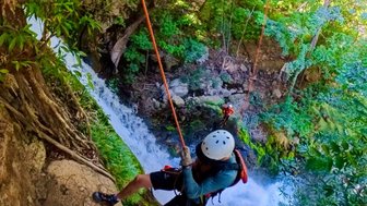 Ein klimmender Abseiler hängt an einem Seil und nähert sich einem Wasserfall in üppig grüner Umgebung.