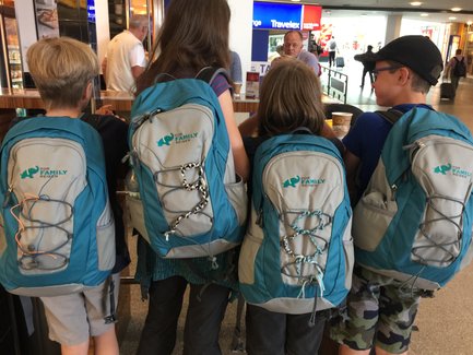 Vier Kinder stehen in einer Schlange, ihre Rucksäcke mit bunten Schnüren sind gut sichtbar und tragen das Logo 'FOR FAMILY REISEN'.