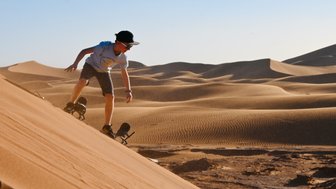 Kind gleitet mit einem Sandboard die sandigen Dünen der Wüste hinunter – Familienreise nach Marokko