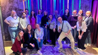 Eine Gruppe von 17 Personen posiert fröhlich vor einer bunten Wand mit leuchtenden Streifen und trägt orangefarbene Brillen.