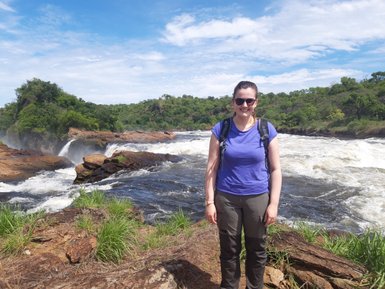 Eine Frau steht lächelnd am Ufer des Murchison Falls, umgeben von üppiger Natur und sprudelndem Wasser.