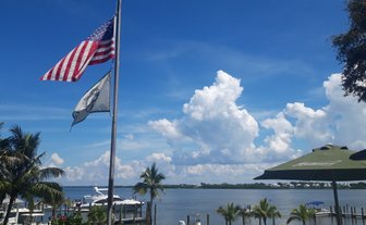 Eine amerikanische Flagge weht über einem malerischen Hafen mit Booten und üppigen Palmen im Vordergrund.