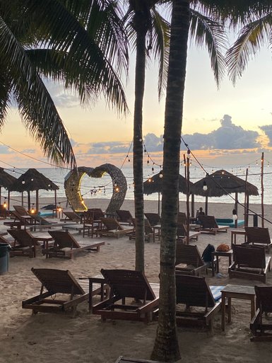 Ein romantischer Strand mit Liegen, Palmen und einem herzförmigen Lichtobjekt, das den Sonnenuntergang beleuchtet.