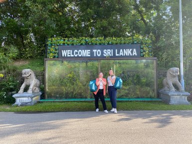 Zwei Frauen machen ein Erinnerungsfoto vor dem „Welcome to Sri Lanka“-Schriftzug am Flughafen – Sri Lanka Reise mit Kindern