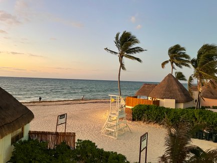 Ein ruhiger Strand mit sanften Wellen, umgeben von Palmen und traditionellen Strohdächern, während die Sonne untergeht.